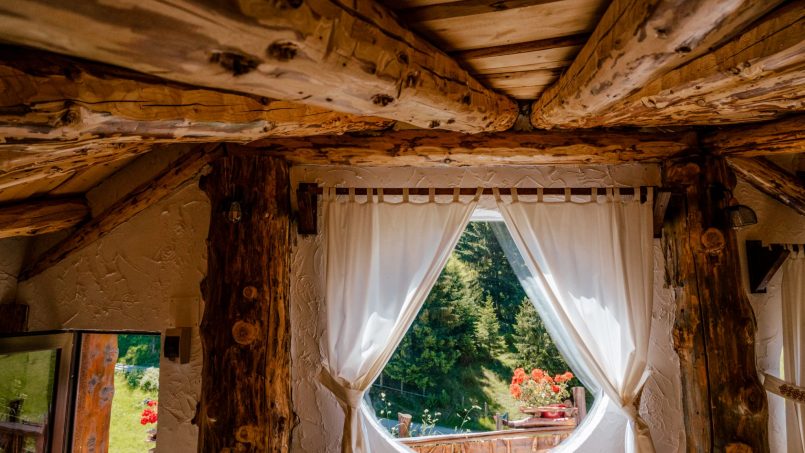 Camera rustică cu fereastra rotundă, tavan din lemn și vedere către natură