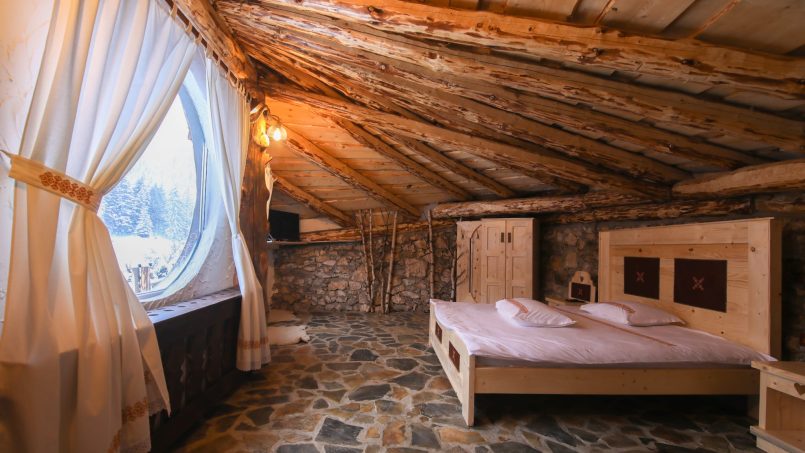 Camera rustică cu pat dublu, fereastra rotundă, podea din piatră și vedere către natură