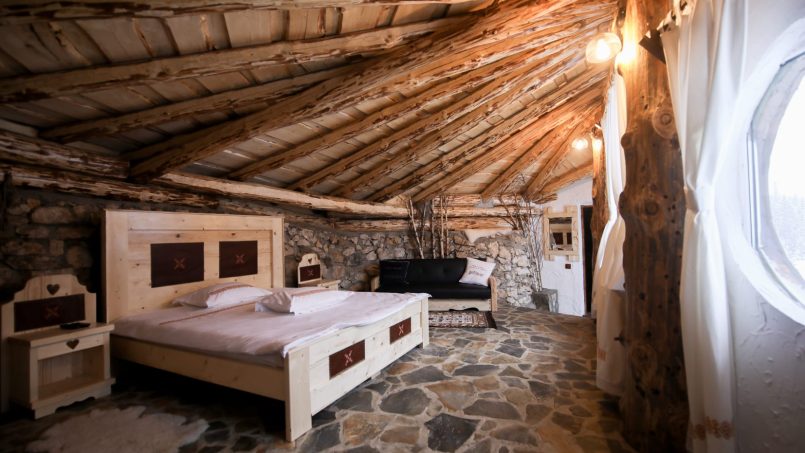 Camera rustică confortabilă cu pat dublu, pereti din piatră, podea de pietre naturale, tavan din lemn și vedere către fereastra rotundă