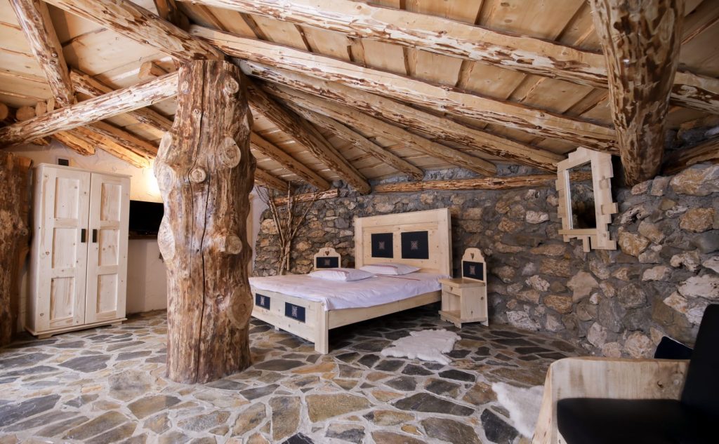 O cameră rustică cu arhitectură naturală, caracterizată prin pereți din piatră, podea de pietre naturale, un pat simplu cu draperii bordate florale, un covor ornamentat cu motive tradiționale și elemente de lemn sculptat care adaugă o atmosferă calda