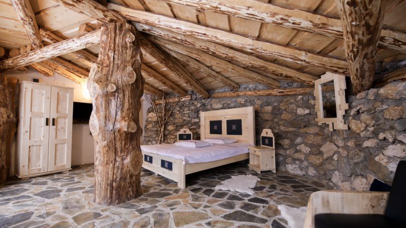 O cameră rustică cu arhitectură naturală, caracterizată prin pereți din piatră, podea de pietre naturale, un pat simplu cu draperii bordate florale, un covor ornamentat cu motive tradiționale și elemente de lemn sculptat care adaugă o atmosferă calda